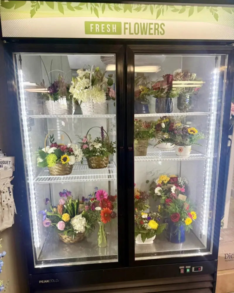 A double-door commercial refrigerator with glass doors, labeled 'FRESH FLOWERS' at the top, filled with assorted bouquets and flower baskets on white shelves. The unit appears to be a Prowl model, with branding visible at the bottom left.