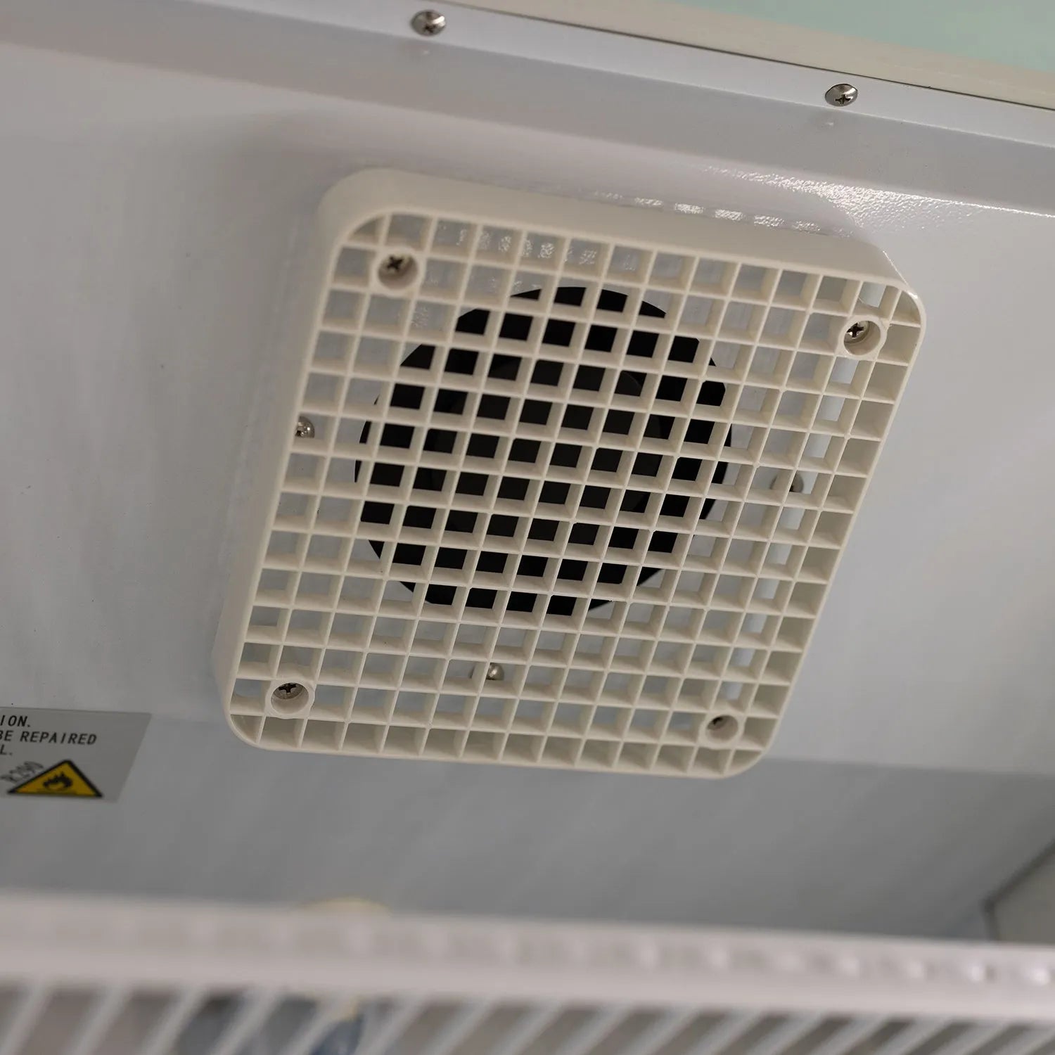 Grid-patterned air vent on a white surface with a warning label.