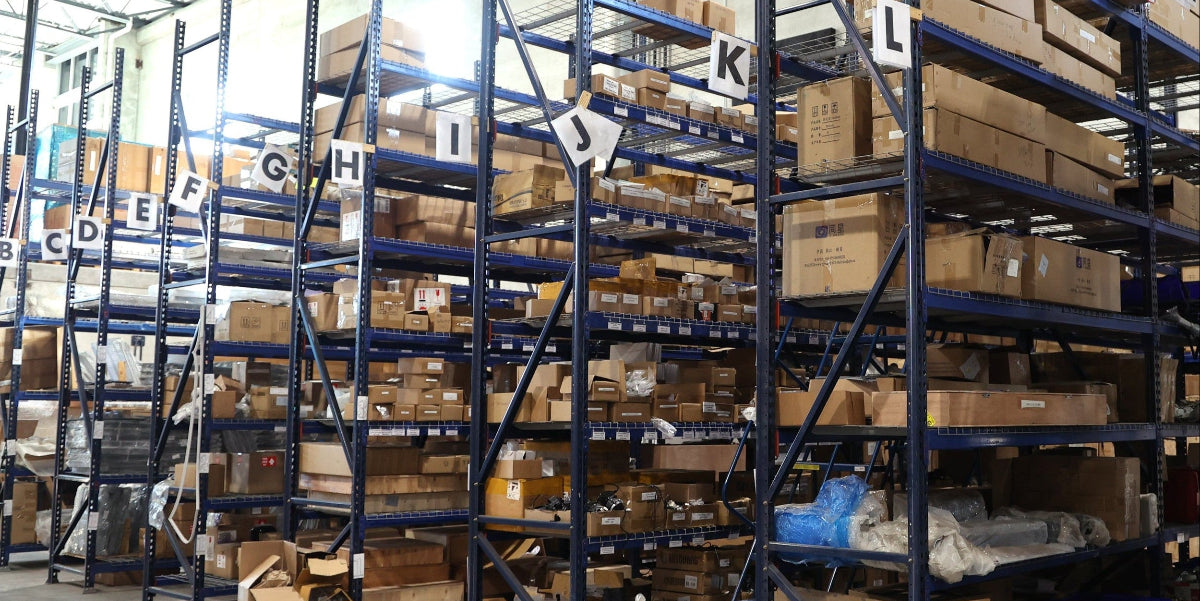 A warehouse interior with tall blue metal shelving units filled with brown cardboard boxes, under a high ceiling with visible structural beams and bright overhead lights.